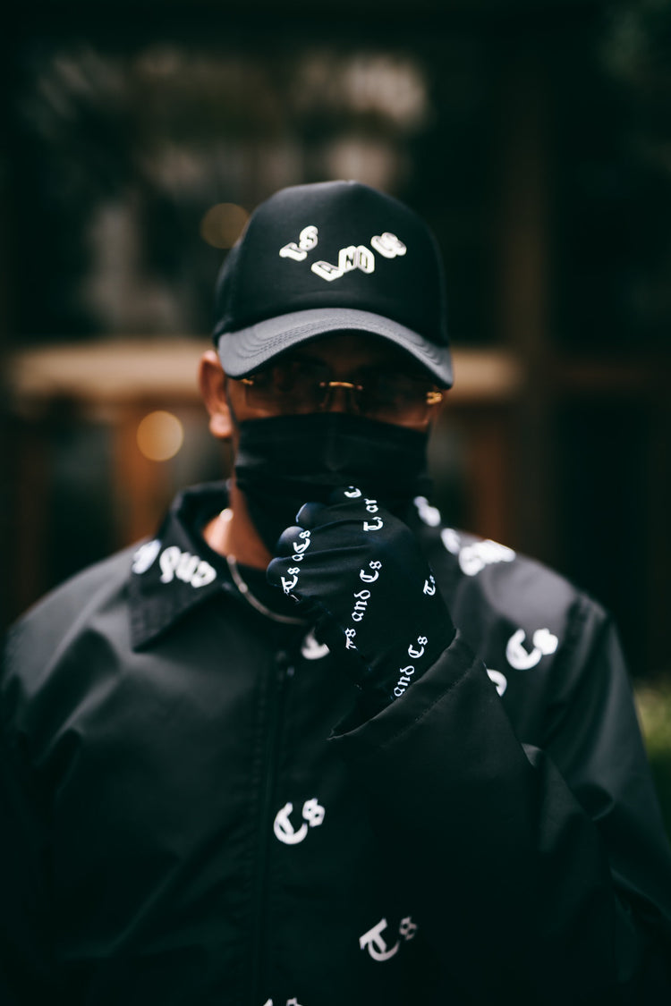 Model in a black Ts and Cs all-over print coach jacket, cap and face covering